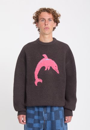 Jeune homme aux cheveux bouclés portant un pull marron foncé avec un grand motif de dauphin rose, associé à un pantalon à carreaux bleu.