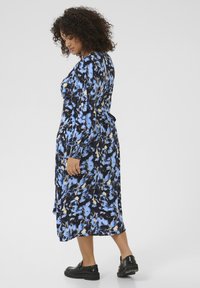 Robe longue avec un motif floral en bleu, noir et crème, avec des manches longues et une taille nouée, associée à des chaussures noires.