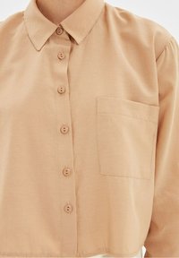 Camicia button-up marrone chiaro realizzata in tessuto morbido, con colletto, sei bottoni e una tasca sul petto con cuciture precise.