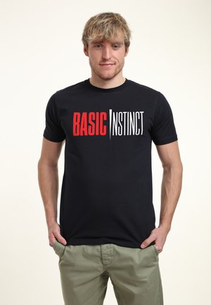 Schwarzes T-Shirt mit dem Text "BASIC" in Rot und "INSTINCT" in Weiß, Rundhalsausschnitt, slim fit, kombiniert mit khakifarbenen Hosen.