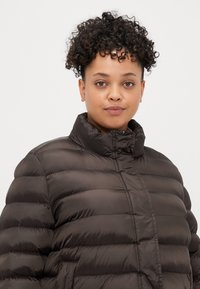 Rjav napihljiv jopič s horizontalnim šivanjem, visokim ovratnikom in sprednjim zadrgo. Izdelan iz sijočega, lahkega materiala. Idealen za toploto.