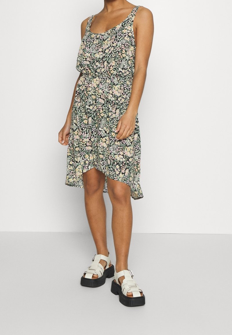 Femme portant une robe florale sans manches et des sandales blanches à plateforme, debout contre un mur clair uni.