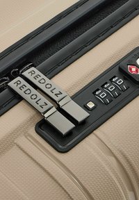 Maleta dura beige con un acabado texturizado, que cuenta con cremalleras negras, un candado de combinación y detalles metálicos con "REDOLZ" grabado.