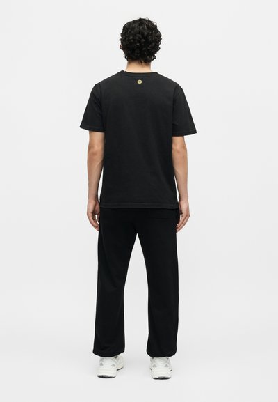 Άνδρας με σγουρά μαλλιά στέκεται γυρισμένος με την πλάτη, φοράει μαύρο μπλουζάκι, μαύρο παντελόνι και λευκά αθλητικά παπούτσια μπροστά σε απλό ανοιχτόχρωμο φόντο.