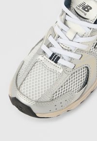 Stříbrná běžecká bota s designem z prodyšné síťoviny, syntetickými překryvy, bílými tkaničkami a černou gumovou podrážkou s ozdobnými detaily.