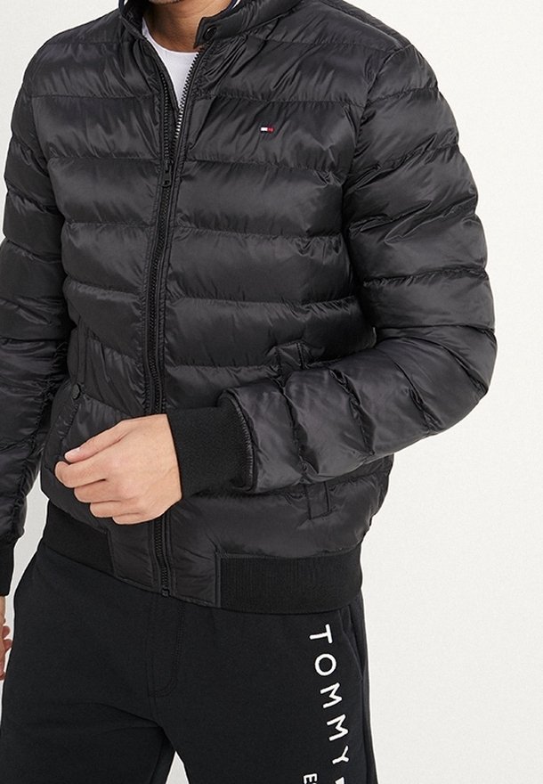 Veste bomber noire rembourrée avec fermeture éclair à l'avant, poignets et taille côtelés. Détail logo discret sur la poitrine et texture lisse et brillante.