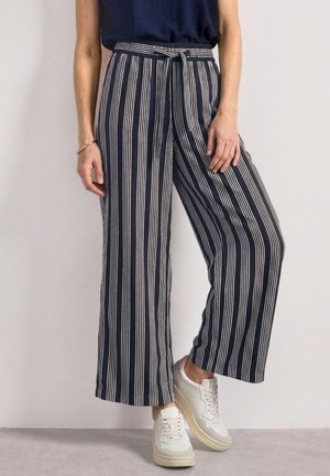 Personne portant un pantalon large rayé verticalement bleu marine et blanc, des baskets blanches et un haut sombre sur un fond clair.
