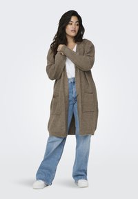 Lång, öppen kort cardigan i en strukturerad brun stickning, med två framfickor, bärs över en vit skjorta och blå vida jeans.