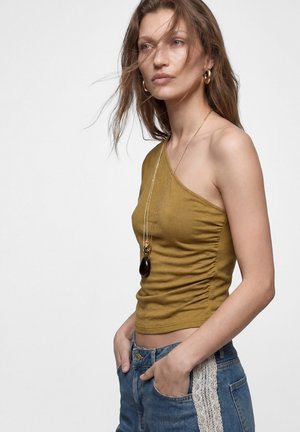 Kvinde med langt hår bærer en sennepsgul ens skulder top, blå jeans med hvidt blondebånd, guld øreringe og et langt vedhængshalskæde.