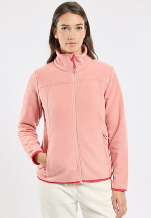 Jeune femme debout portant une veste polaire rose zippée avec bordure rouge et un pantalon blanc sur un fond blanc uni.
