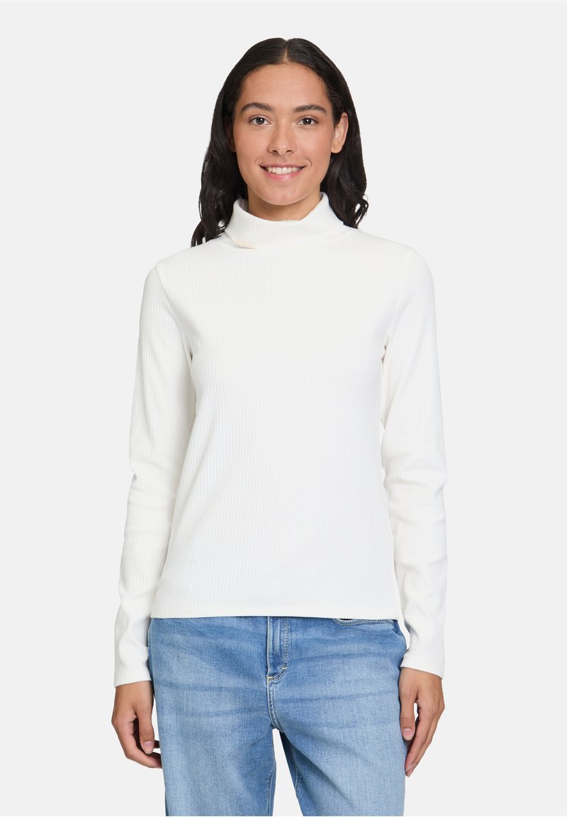 Weißes geripptes Langarm-Turtleneck-Oberteil mit schmaler Silhouette, kombiniert mit hellblauen Jeans. Der Stoff wirkt weich und dehnbar.