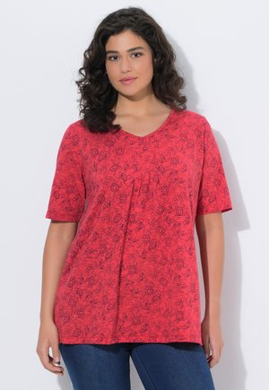 Frau mit lockigem dunklem Haar, die ein rotes Kurzarm-Top mit dezentem Blumenmuster und eine blaue Jeans trägt, vor einem weißen Hintergrund steht.