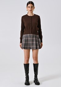 Cardigan marrone a coste con chiusura a bottoni, abbinato a una gonna plissettata scozzese in toni scuri e stivali neri in pelle al ginocchio.