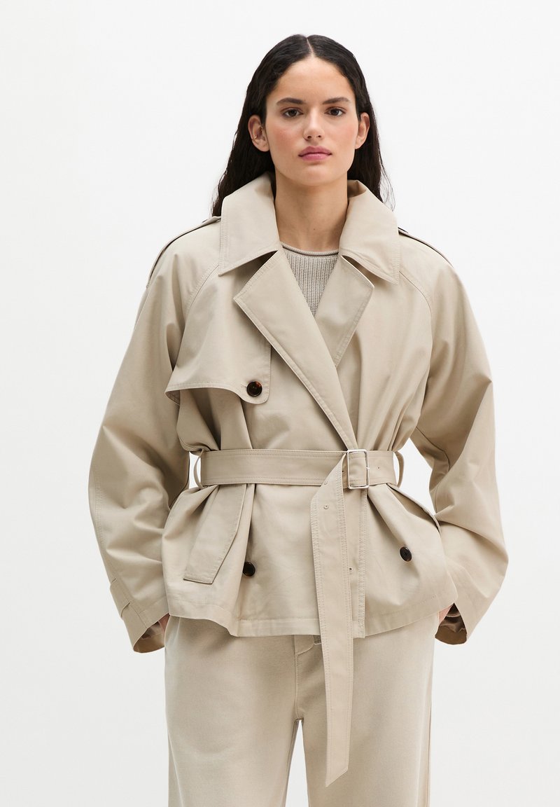 Donna con capelli lunghi e scuri che indossa un trench beige con cintura abbinato a pantaloni coordinati, in piedi contro uno sfondo chiaro e uniforme.