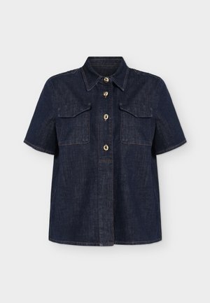 Femme aux cheveux blonds courts portant une chemise sombre à manches courtes, un pantalon beige ample, des chaussures noires et tenant un sac à main noir sur fond blanc.