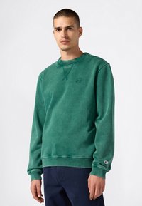 Hombre con un sudadera verde de manga larga con un pequeño logo en el pecho y la muñeca, combinada con pantalones azul oscuro, de pie contra un fondo plain.