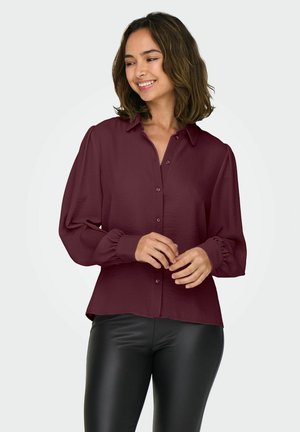 Blouse bordeaux avec un devant à boutons, un col et de longues manches bouffantes. Texture lisse, poignets froncés et coupe décontractée. Associée à des leggings noirs.