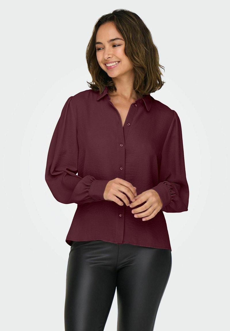 Blouse bordeaux avec un devant à boutons, un col et de longues manches bouffantes. Texture lisse, poignets froncés et coupe décontractée. Associée à des leggings noirs.