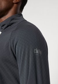 Haut de sport long gris foncé avec un col haut, en tissu texturé et un motif géométrique subtil ; présente un logo sur l'épaule.