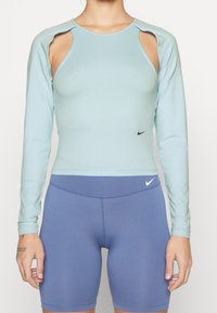 Hellblaues langärmliges Croptop mit ausgeschnittenen Schultern, aus dehnbarem Stoff, kombiniert mit hoch taillierten, helllila Shorts mit Nike-Logos.