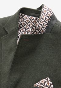 Blazer vert foncé en tissu texturé, avec une doublure intérieure de col à motif dans des tons crème, marron et bordeaux. Comprend un mouchoir de poche.