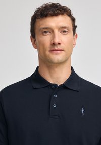 Svart polo t-shirt med ribbad krage, tre knappar framtill och en liten broderad logotyp på vänster bröst. Slät textur och klassisk design.