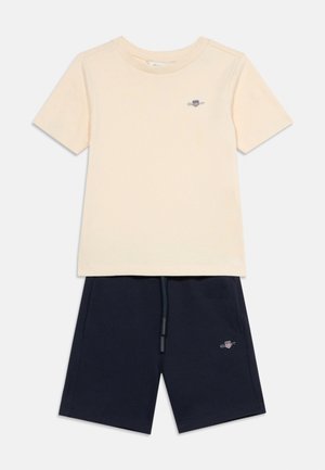 Μπεζ κοντομάνικη μπλούζα και σκούρο μπλε σορτς με κορδόνι, και τα δύο με μικρά κεντημένα λογότυπα στην αριστερή πλευρά.