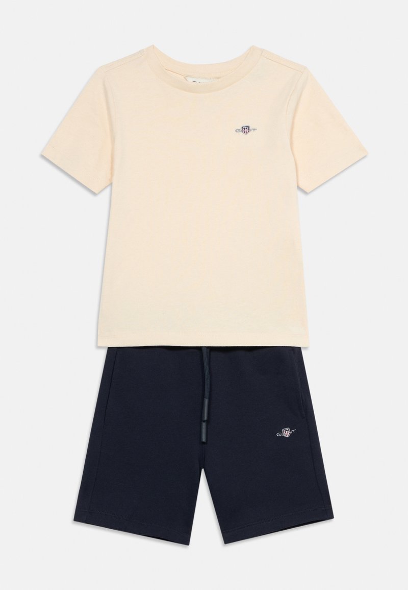 Μπεζ κοντομάνικη μπλούζα και σκούρο μπλε σορτς με κορδόνι, και τα δύο με μικρά κεντημένα λογότυπα στην αριστερή πλευρά.