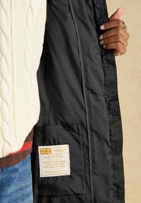 Czarna pikowana kurtka z kieszenią z beżową metką. W pobliżu widoczny sweter w kolorze kremowym z splotem w warkocze. Minimalistyczny design i funkcjonalne elementy.