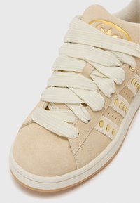 Sapatilhas de camurça bege com atacadores grossos de creme, detalhes em dourado e riscas diagonais de lado. Sola de borracha e suporte acolchoado para o tornozelo.
