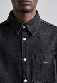 Camicia in denim nero con chiusura a bottoni, colletto a punta, bottoni argentati e taschino sul lato sinistro con la scritta "SANDRO" ricamata in bianco.