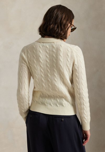 Vrouw met kort bruin haar die schildpadzonnebril en een crème kabelgebreide trui draagt, gecombineerd met een donkere broek, van achteren gezien.