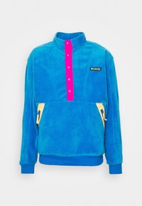 Pullover in pile di colore blu brillante con colletto alto, patta con bottoni rosa, zip delle tasche gialle e una piccola etichetta con logo.