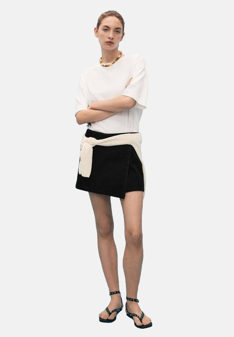 Bela majica s kratkimi rokavi, črn mini jeans krilo s kremasto jopico zavezano okoli pasu in črne sandale z naramnicami ter okrasnimi kovinskimi dodatki.