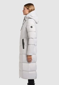 Long, light gray puffer coat with a hood, featuring a quilted design, black hardware accents, and a zippered side pocket.