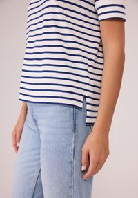 Weiß und dunkelblau gestreiftes T-Shirt mit kurzen Ärmeln und seitlichen Schlitzen am Saum, kombiniert mit hellblauen Jeans. Glatte Stoffstruktur.