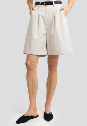 Kreminės lininės šortai su klostėmis, apvartu ir šoninėmis kišenėmis. Nešiojami su juodu diržu ir juodais slip-on batais.