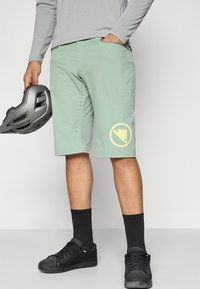 Svijetlozelene biciklističke kratke hlače s perforiranim dizajnom duž bočnih strana i žutim logotipom na lijevoj nozi, u paru s crnim čarapama i cipelama.