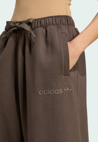 Osoba s smeškim smeškom nosi smeđu Adidas trenirku s remenom, ruku u bočnom džepu, a Adidas logo vidljiv na butu.