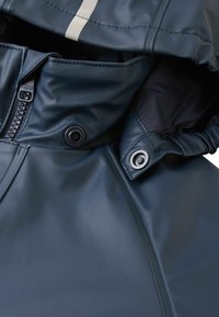 Granatowa wodoodporna kurtka z zapięciem na zamek, zatrzaskami, elastycznymi mankietami oraz odblaskowymi paskami, o gładkiej, błyszczącej fakturze.