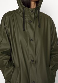 Giacca verde oliva impermeabile con cappuccio, bottoni a pressione e coulisse. Progettata con maniche lunghe e tasche laterali. Tessuto dalla texture liscia.