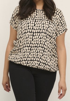 Woman wearing a short-sleeve beige and black patterned blouse paired with black fitted pants, standing against a plain background.