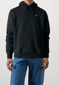 Felpa nera con cappuccio, tasca frontale, coulisse regolabile e piccolo logo sul petto; abbinata a jeans cargo azzurri chiari.