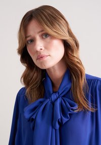 Blusa di chiffon blu con un ampio collo a fiocco, caratterizzata da un tessuto morbido e fluido e da pieghe delicate. Il design è elegante con una texture liscia.