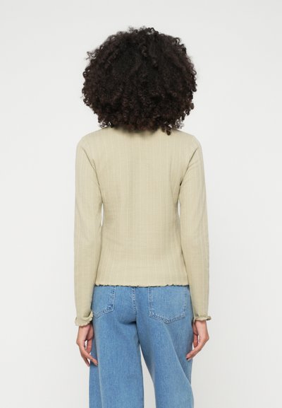 Ανοιχτό πράσινο μακρυμάνικο μπλουζάκι με ριγωτή υφή και στενή εφαρμογή. Το τελείωμα και οι μανσέτες διαθέτουν μια μαλακή, φουσκωτή άκρη. Συνδυάζεται με μπλέ παντελόνι με φαρδιά πόδια.