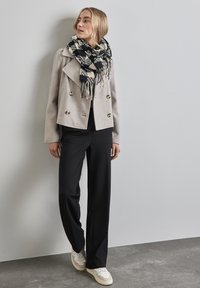 Kvinna iklädd beige kort jacka, svart och vit rutigt scarf, svarta byxor och vita sneakers, stående mot en grå vägg.