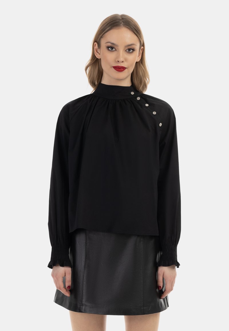 Schwarze Bluse mit hohem Kragen, gerafftem Stoff und langen Ärmeln. Verfügt über dekorative Knöpfe an der Schulter. Kombiniert mit einem schwarzen Lederrock.