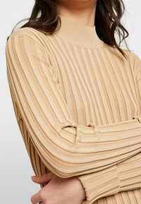 Femme portant un pull beige côtelé à rayures verticales, les bras croisés, montrant une partie de son cou et de ses cheveux.