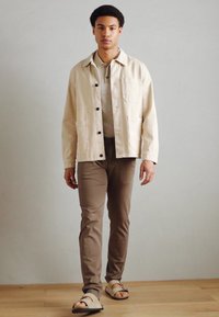 Beige leichte Jacke mit dunklen Knöpfen, über einem hellgrauen gestreiften Hemd und beigen Hosen getragen, ergänzt durch braune Sandalen.