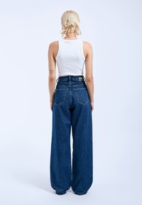 Weiße ärmellose Bluse kombiniert mit hoch taillierten, weiten, dunklen Jeans. Verfügt über zwei Gesäßtaschen und ein Etikett am Bund.